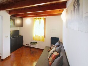 Ein Wohnzimmer mit einer Couch, einem kleinen Tisch, einem Fenster mit Vorhängen und einer Küchenzeile mit Schränken.