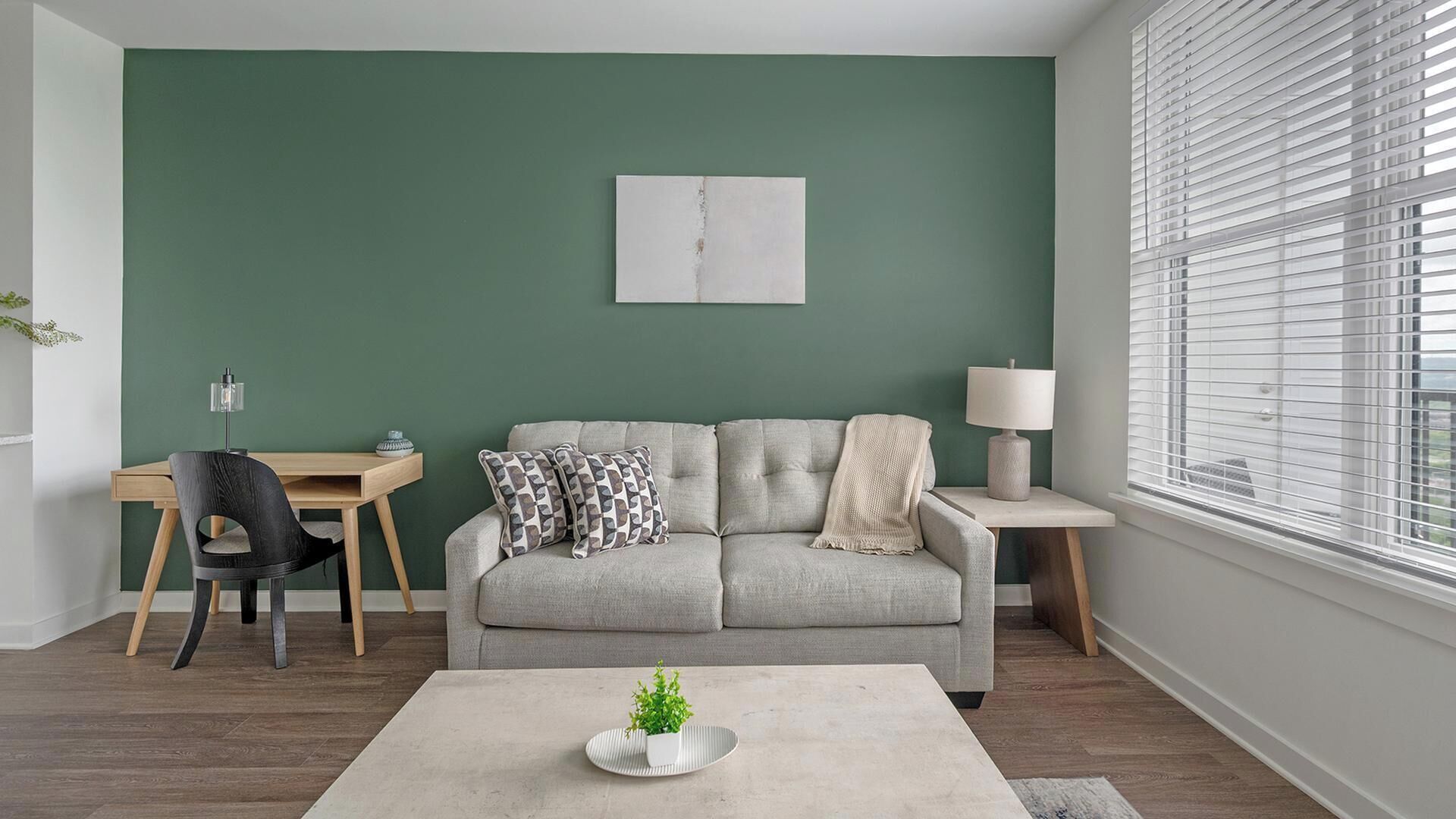 Una sala de estar con pared verde, un sofá gris, una mesita, una lámpara y un ventanal con persianas.