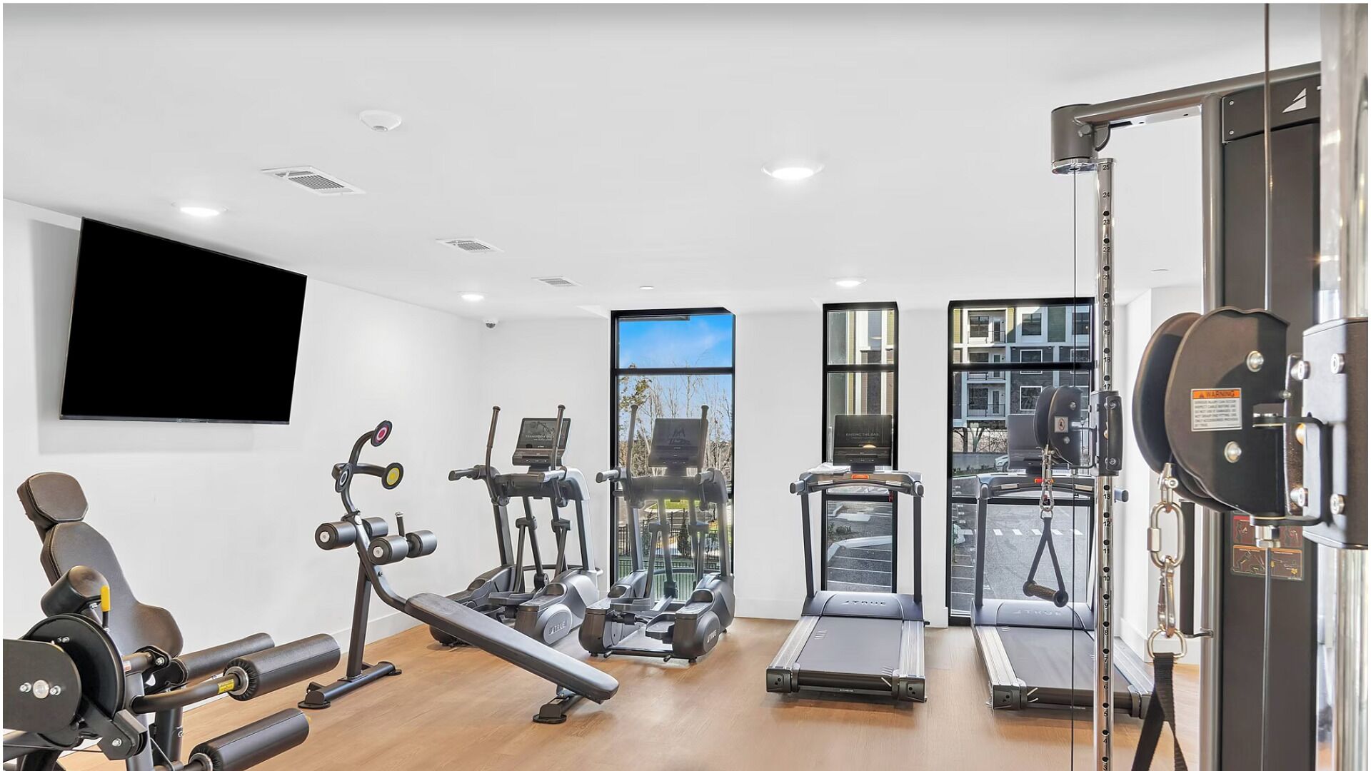 A modern gym with cardio machines, weightlifting equipment, and a large window offering a city view.