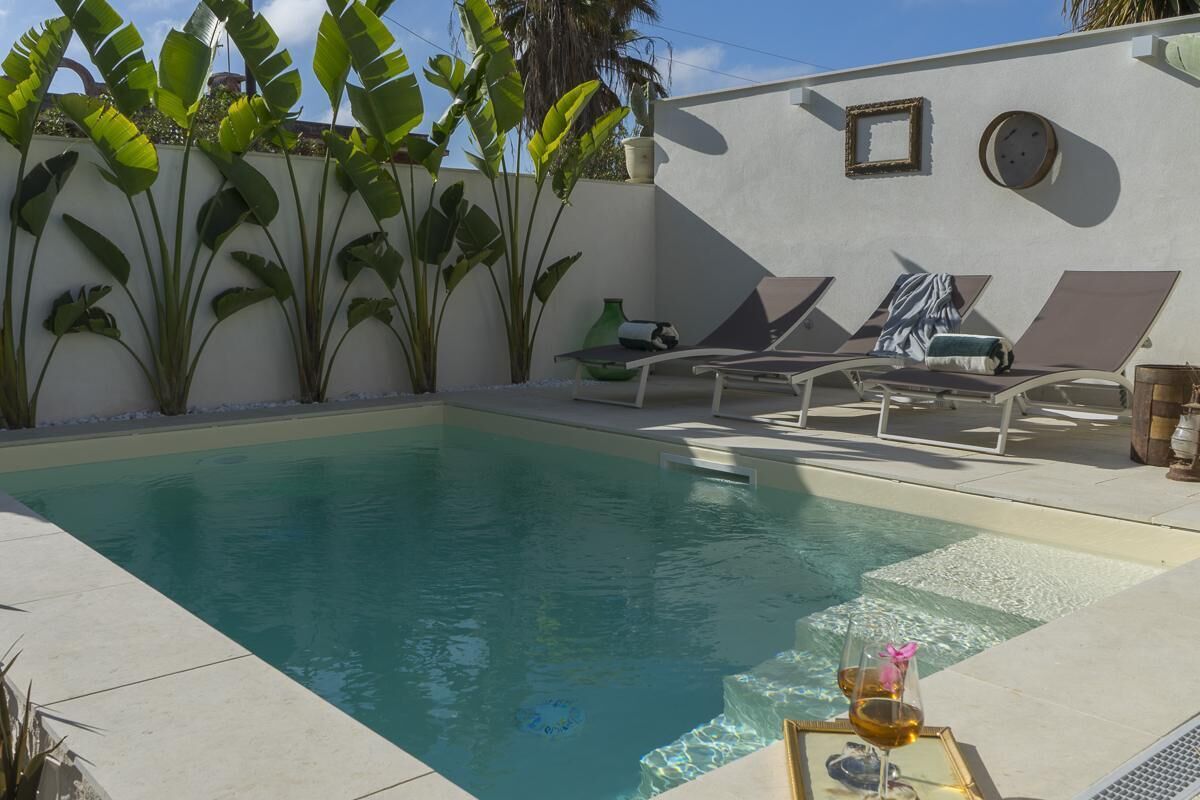 A swimming pool with lounge chairs, a glass table with a drink, and tropical plants.
