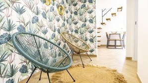 Una habitación con papel tapiz estampado con hojas tropicales, dos sillones de mimbre, un tapete tejido y un comedor con mesa y sillas.