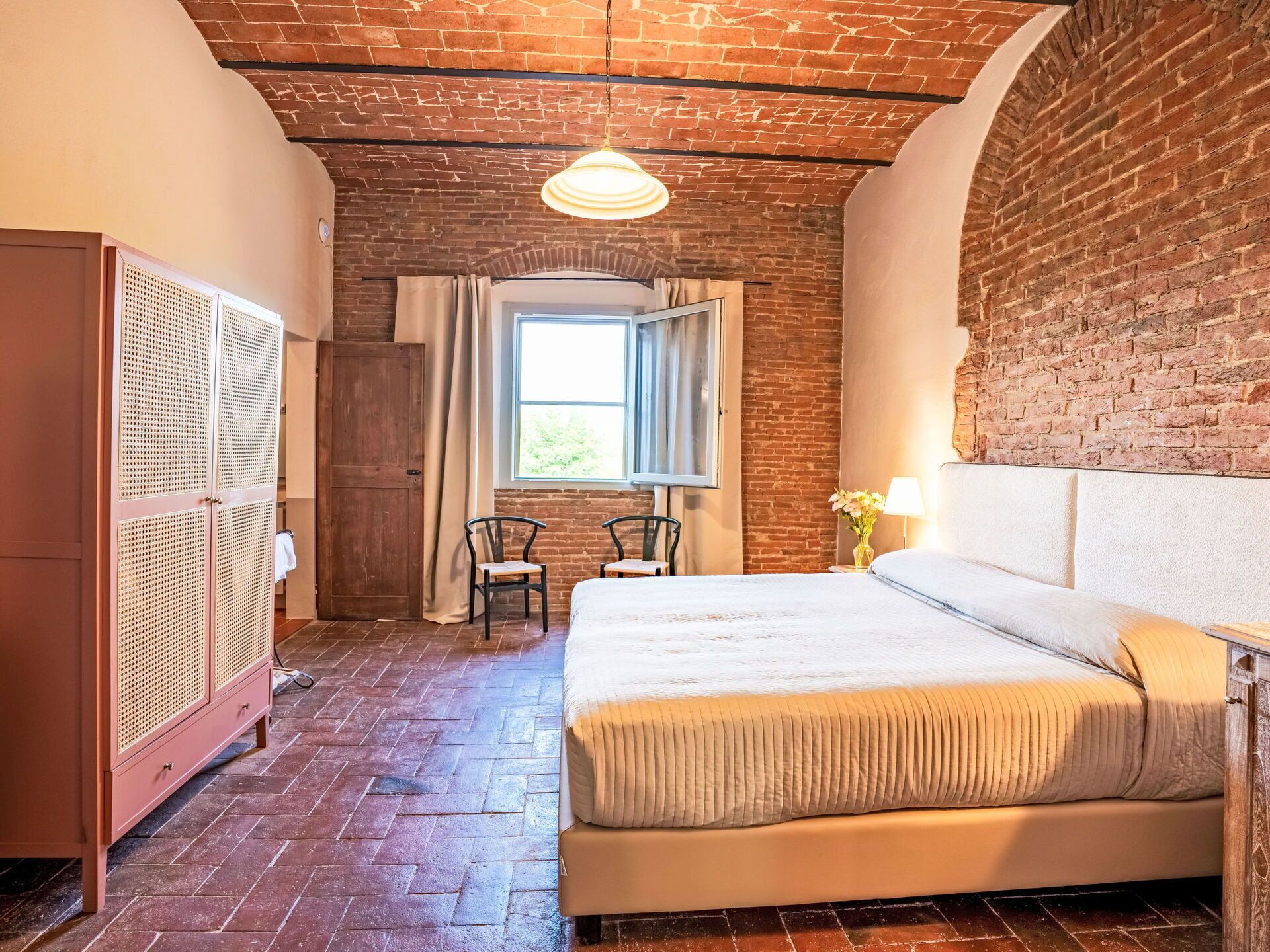 Une chambre avec un mur en briques, une armoire en bois, un lit avec une tête de lit blanche, deux chaises et une fenêtre avec des rideaux.