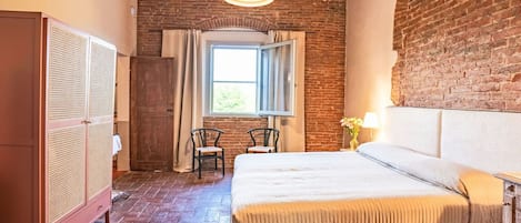 A bedroom with a brick wall, a wooden wardrobe, a bed with a white headboard, two chairs, and a window with curtains.