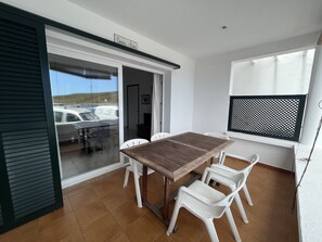 Un balcon avec une table en bois et des chaises en plastique blanc, une porte-fenêtre coulissante et une fenêtre ornée d’un treillis noir.