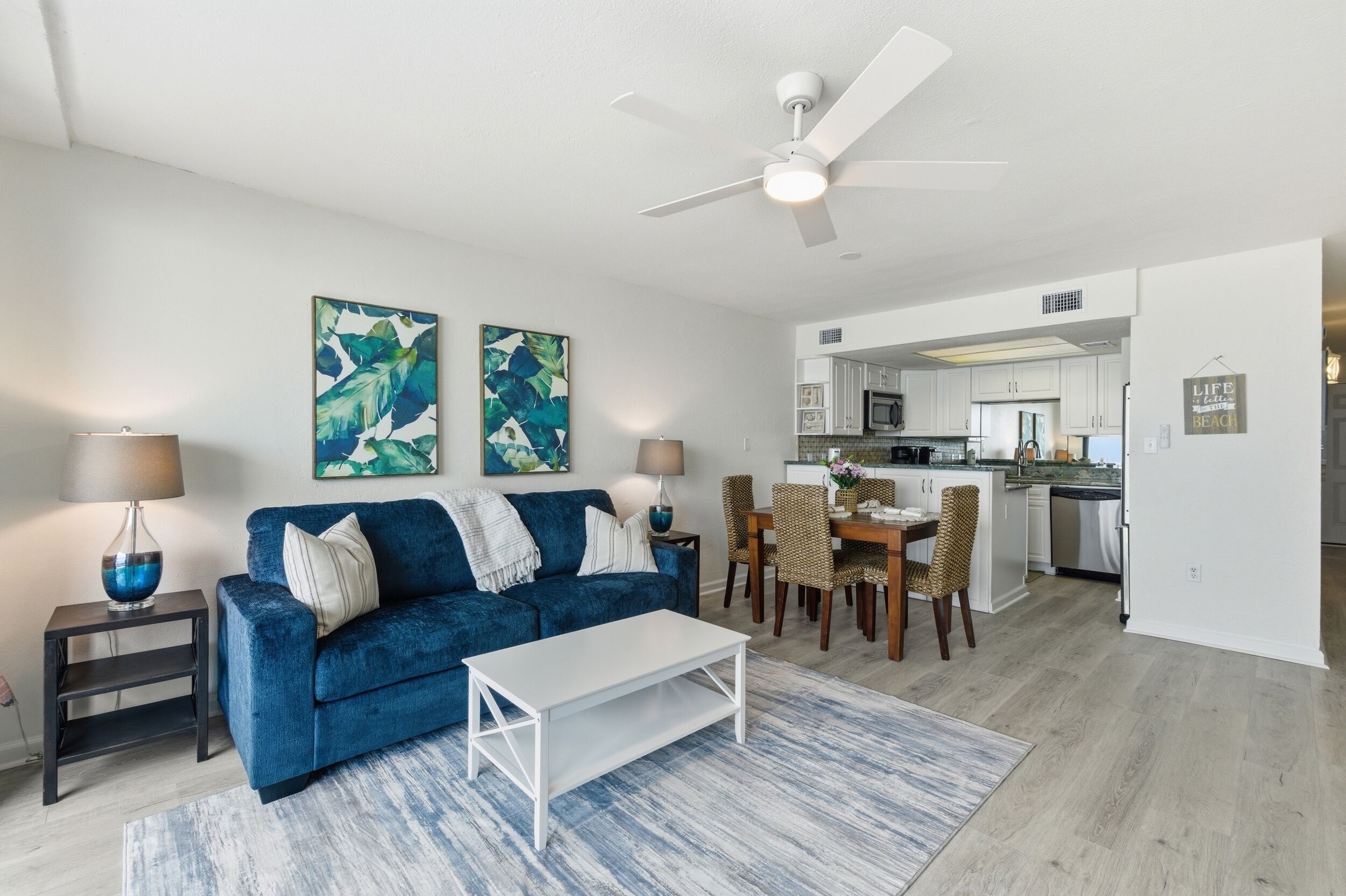 A living room with a blue sofa, a white coffee table, and a dining area with wicker chairs.