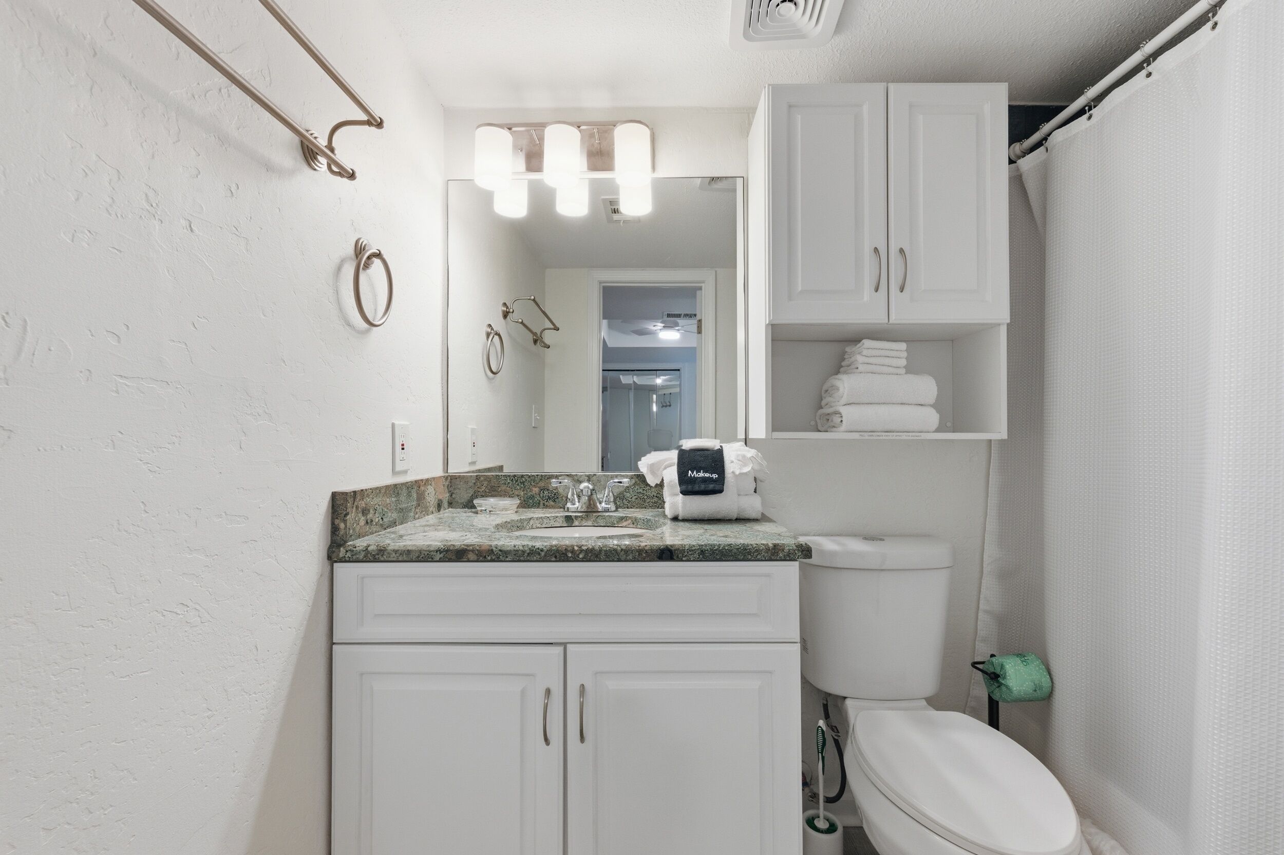 Une salle de bain avec des toilettes blanches, un lavabo doté d’un plan de travail en marbre, un porte-serviettes et un rideau de douche.