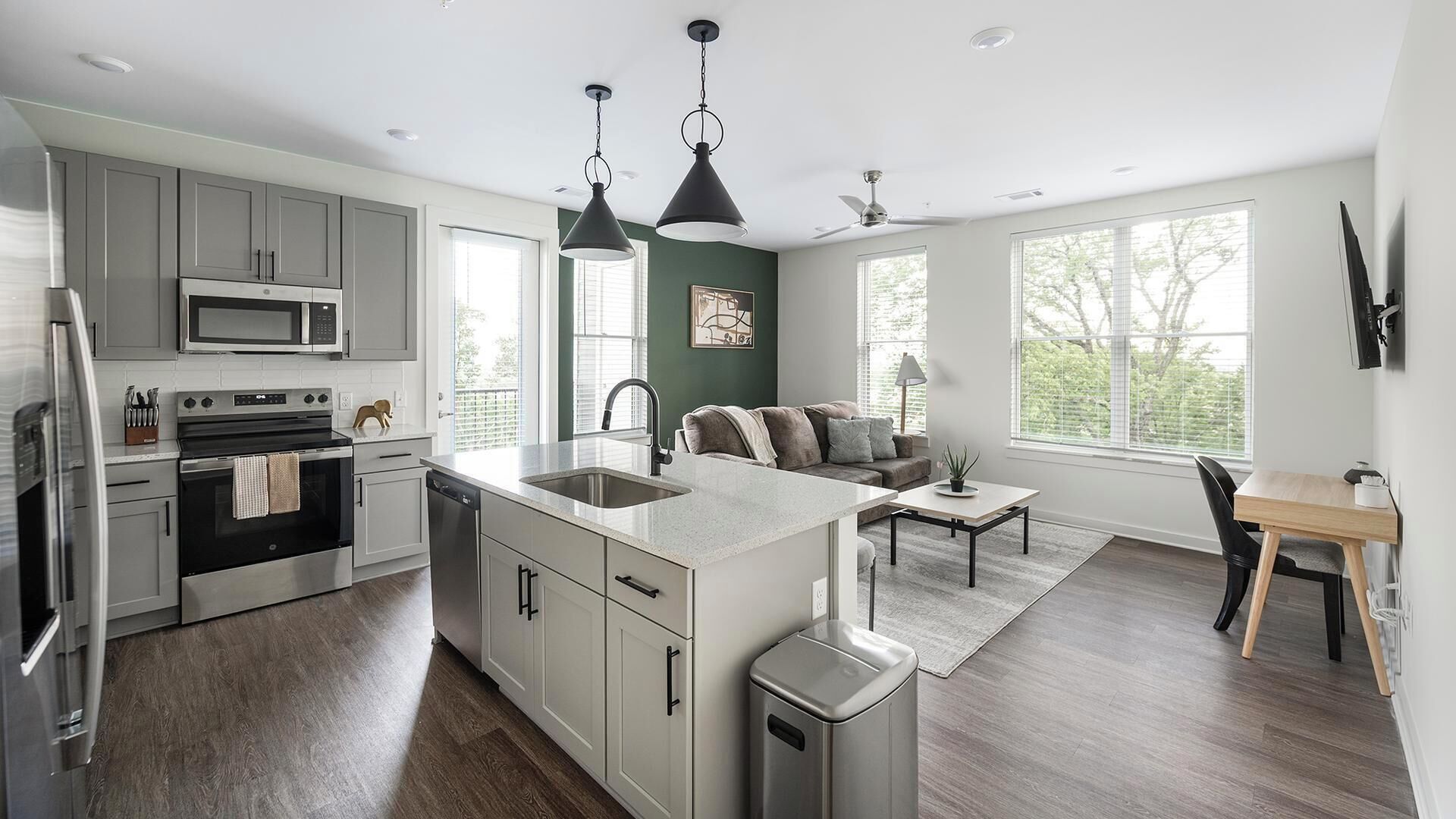 A modern kitchen with grey cabinets, a microwave, and a dishwasher. There's a dining area with a table and chairs, a sofa, and a ceiling fan.