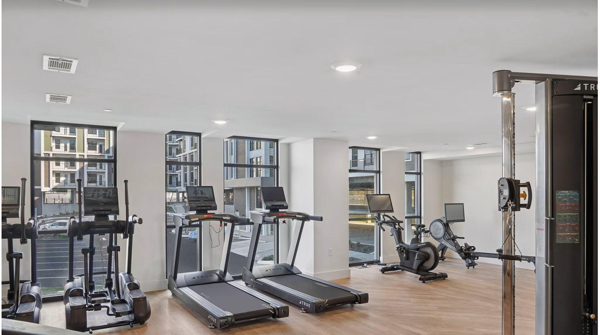 A modern gym with treadmills, elliptical machines, and a rowing machine, large windows, and a view of buildings outside.