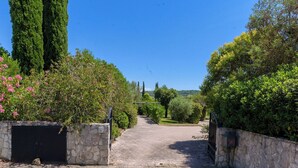 Un camino empedrado que conduce a una entrada con reja, rodeado de abundante vegetación y muros de piedra.