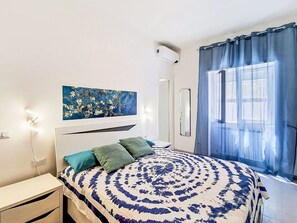 Un dormitorio con cama, mesita de noche, aire acondicionado instalado en la pared, ventana con cortinas y un cuadro en la pared.