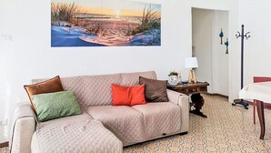 Ein Wohnzimmer mit einem großen Wandbild, einer Couch mit Kissen, einem kleinen Tisch mit Lampe und einem Kleiderständer.