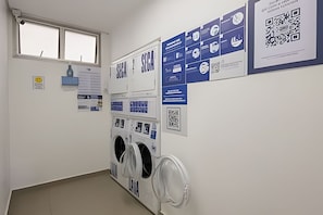 Uma lavanderia com máquina de lavar e secadora fixadas na parede, uma janela e diversos cartazes informativos.