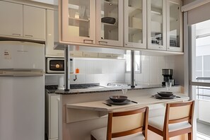 A modern kitchen with white cabinetry, a built-in oven, a coffee machine, and a dining area with two chairs.