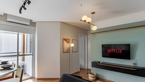 A modern living room with a dining area, a mounted television displaying 'NETFLIX,' and a large window providing natural light. - Coberturinha Massa (São Paulo)