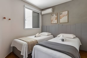 A hotel room with two beds, a window with blinds, an air conditioning unit, and two wall-mounted artworks.