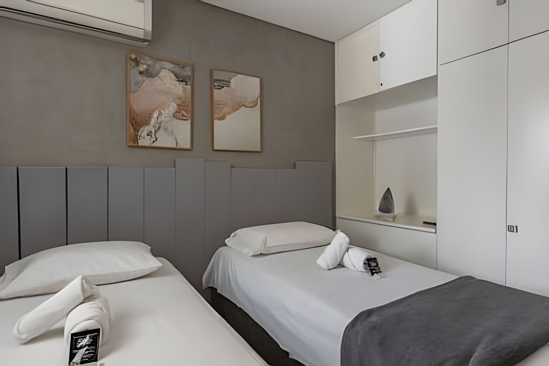 Une chambre d’hôtel avec deux lits, une tête de lit grise, une petite étagère ornée d’un objet décoratif et deux œuvres d’art encadrées accrochées au mur.