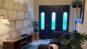 A cozy living room with a stone wall, a dark door with illuminated windows, a wooden console table, a black sofa, and a potted plant.