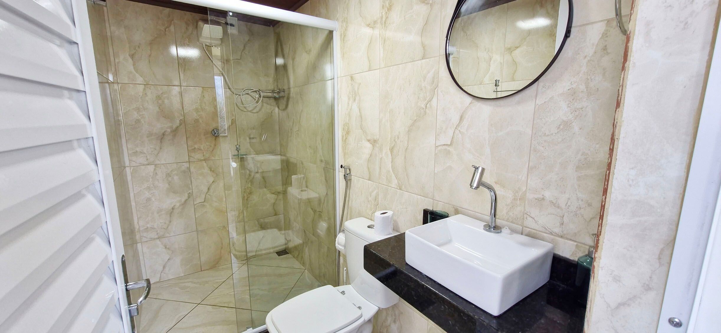Un baño con cabina de ducha de vidrio, un inodoro blanco, un lavabo con encimera negra y un espejo redondo.