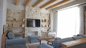 A living room with a stone wall, wooden beams, a flat-screen TV, and a sofa. - Village Escape House by CorfuEscapes (Corfu)