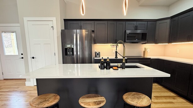 A modern kitchen with dark cabinetry, stainless steel appliances, a white island with stools, and a view of a wooden floor. - 2 King beds! Sleep like royalty in this cozy, intimate, and brand-new house (Scottsville)