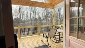 A wooden deck with a rocking chair and a table, a glass door, and a view of trees. - 2 King beds! Sleep like royalty in this cozy, intimate, and brand-new house (Scottsville)