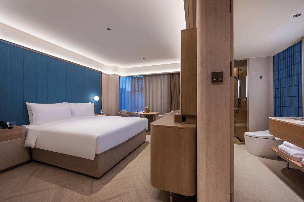 A modern hotel room with a large bed, a desk, a bathroom with a shower, and a view of the city.