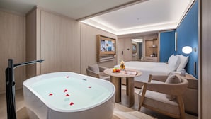 Una habitación de hotel moderna con una bañera grande, una cama, una mesita con fruta y un televisor.