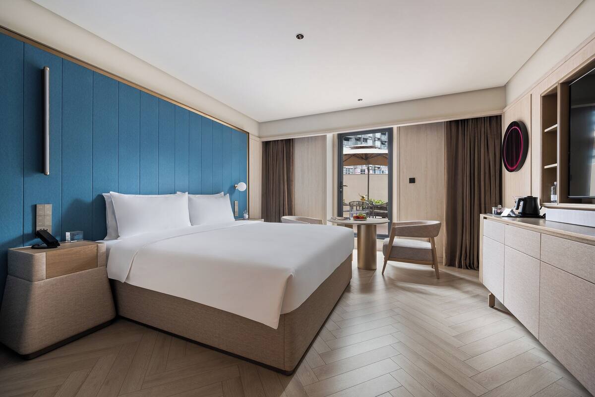 A modern hotel room with a large bed, a desk, a chair, and a view of the city.