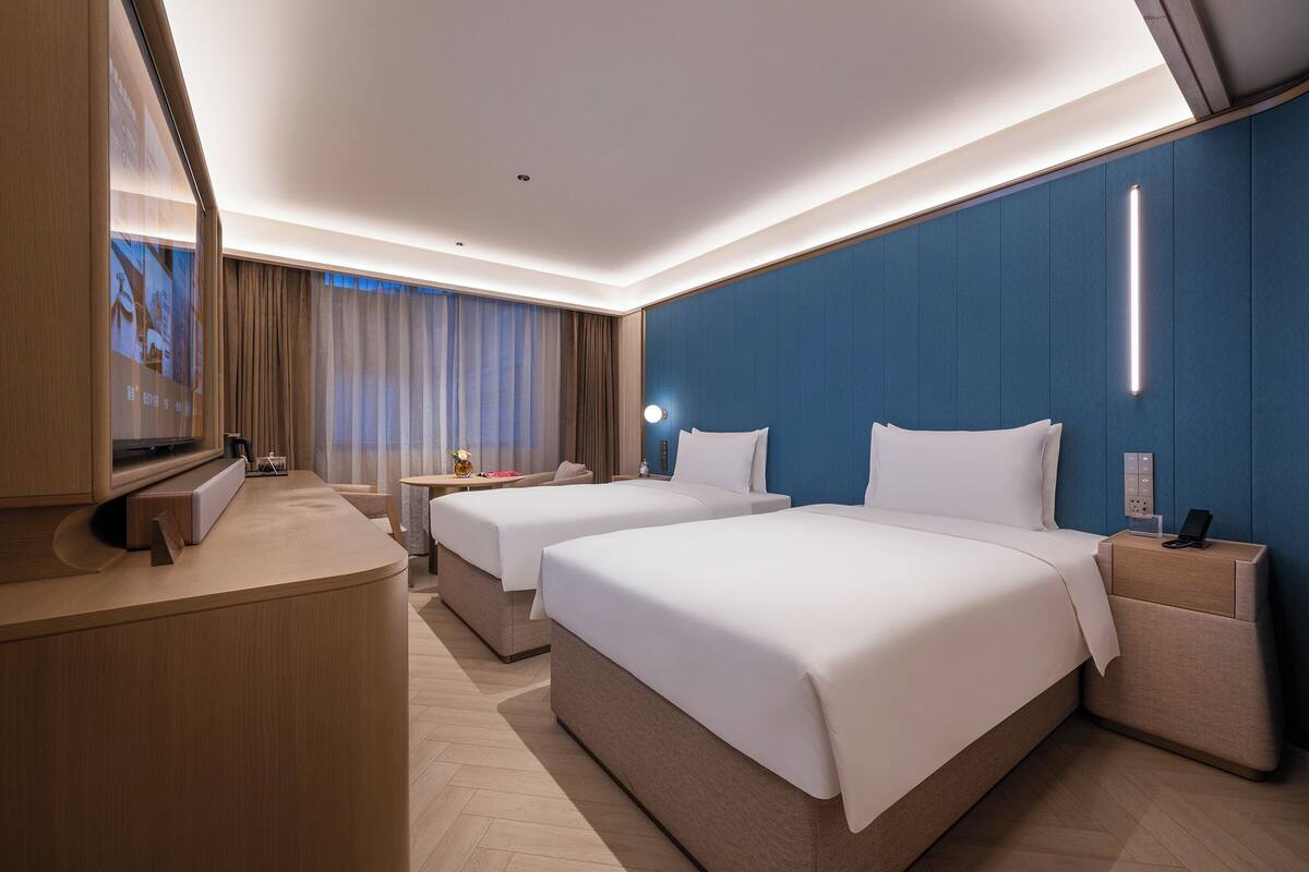 A hotel room with two beds, a desk, a chair, a TV, and a large window with curtains.