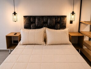 A bed with a quilted headboard, two bedside tables with lamps, and a remote control on one of the tables. - Stay 32 (Medellín)