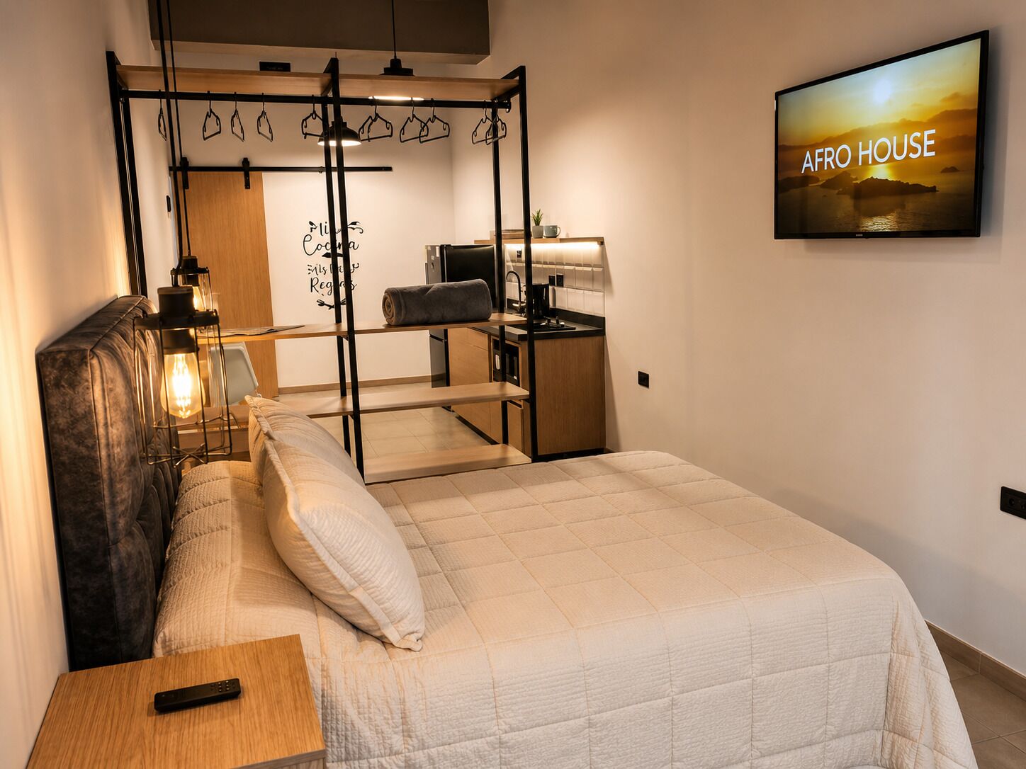 Ein Schlafzimmer mit Bett, Nachttisch, einem Fernseher, der 'AFRO HOUSE' zeigt, einer Küchenzeile und einem Kleiderschrank.