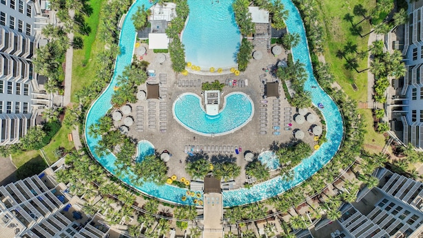 An aerial view of a hotel with a large central pool shaped like a guitar, surrounded by palm trees and lounge areas. - Destin West Bayside Sandpiper #405 (Fort Walton Beach)
