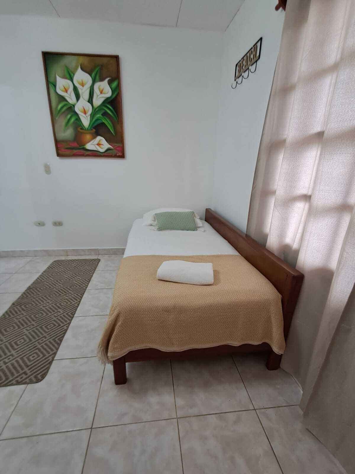 Ein Einzelbett mit brauner Kopfteil, einem weißen Kopfkissen und einer beigen Decke. Darüber an der Wand hängt ein Bild von weißen Blumen.