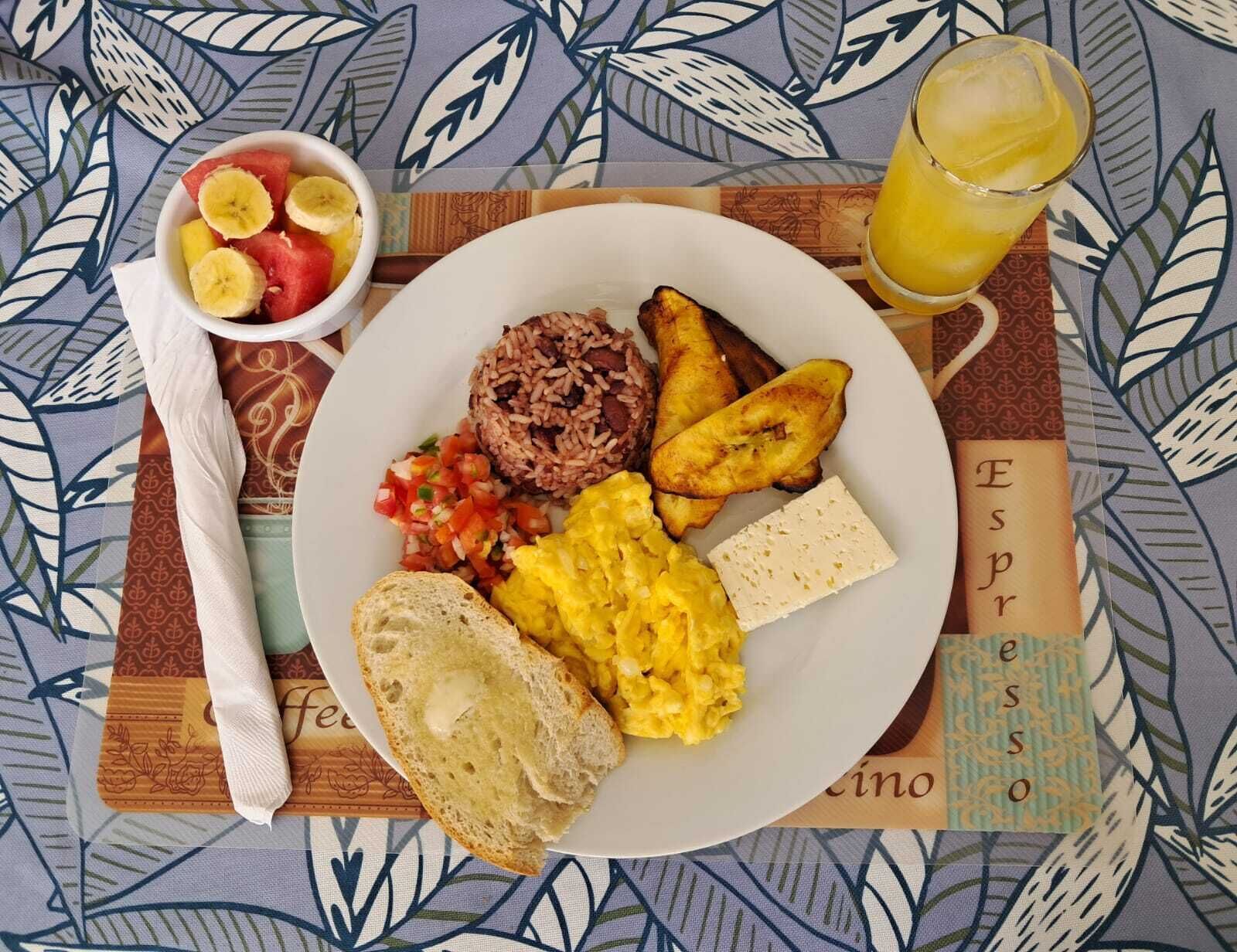 A plate of food with rice, eggs, plantains, and toast, accompanied by a glass of juice and a bowl of fruit.