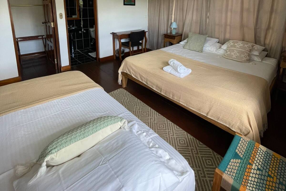 Une chambre d’hôtel avec deux lits, un ventilateur de plafond, une petite table, une chaise, un miroir et une fenêtre avec des rideaux.