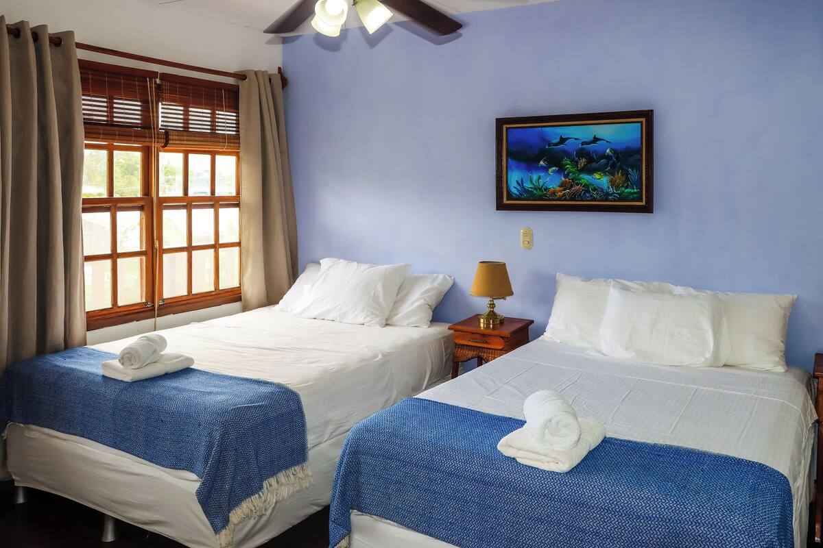 Une chambre d’hôtel avec deux lits, une fenêtre avec des rideaux, un tableau accroché au mur et un ventilateur de plafond.