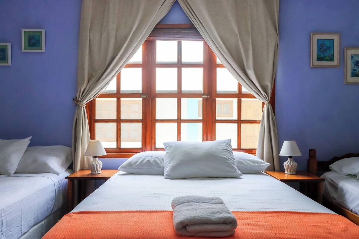 Une chambre d’hôtel avec un grand lit, deux tables de chevet avec des lampes, une fenêtre avec des rideaux et des œuvres d’art accrochées aux murs.