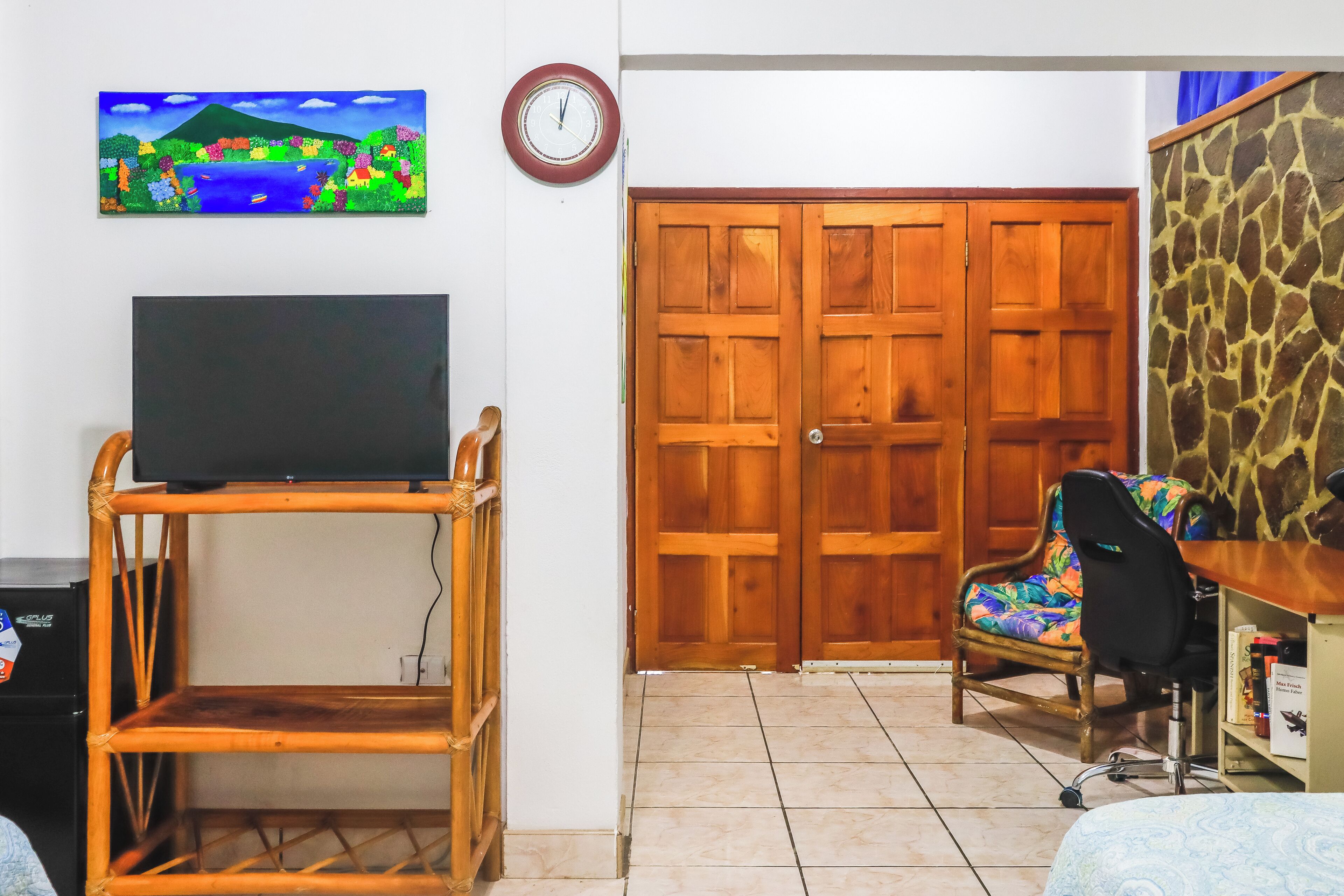 Une pièce avec un téléviseur posé sur un meuble en bois, une porte en bois, une chaise et une horloge murale.