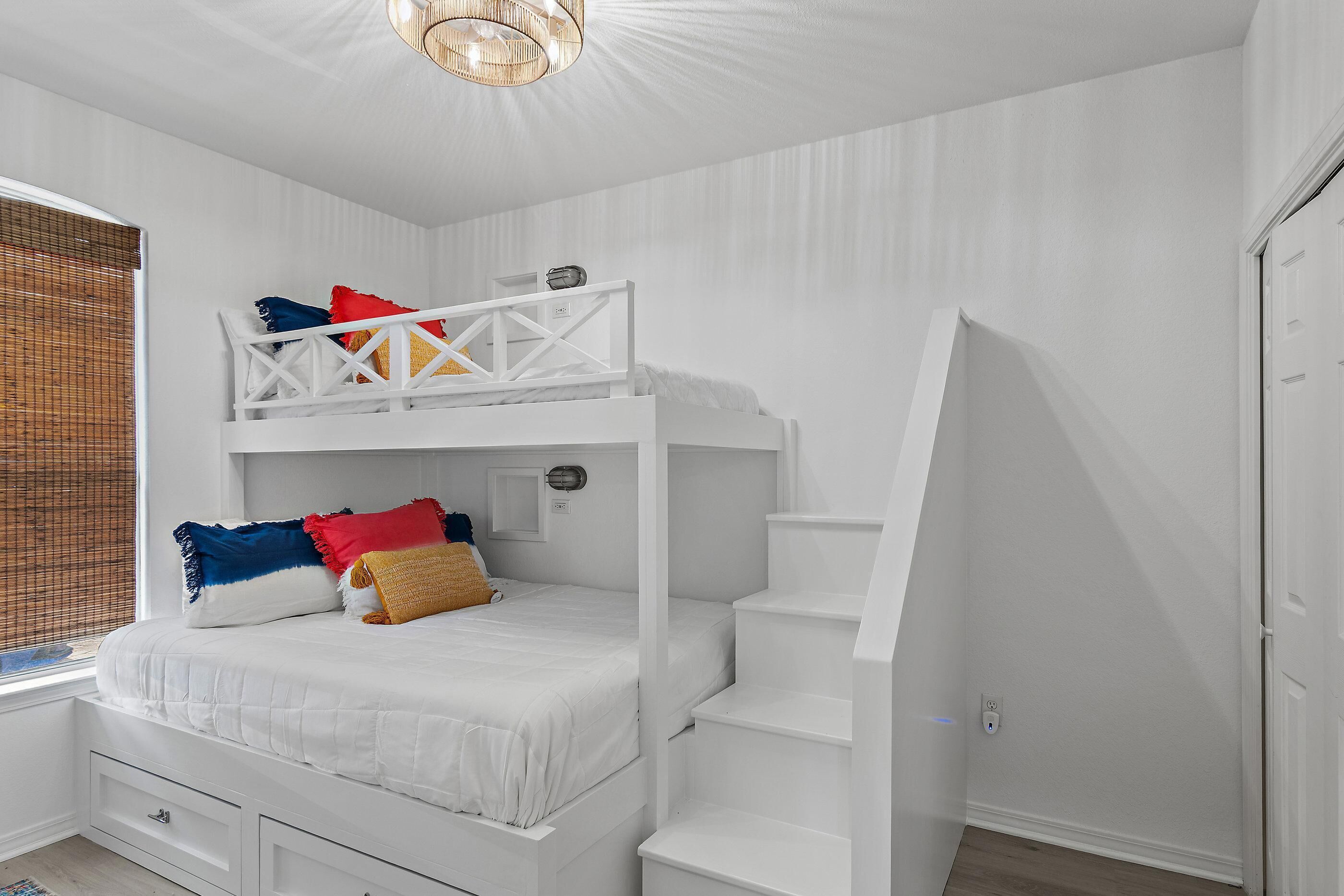 A bunk bed with a white frame, a window with blinds, and a staircase.