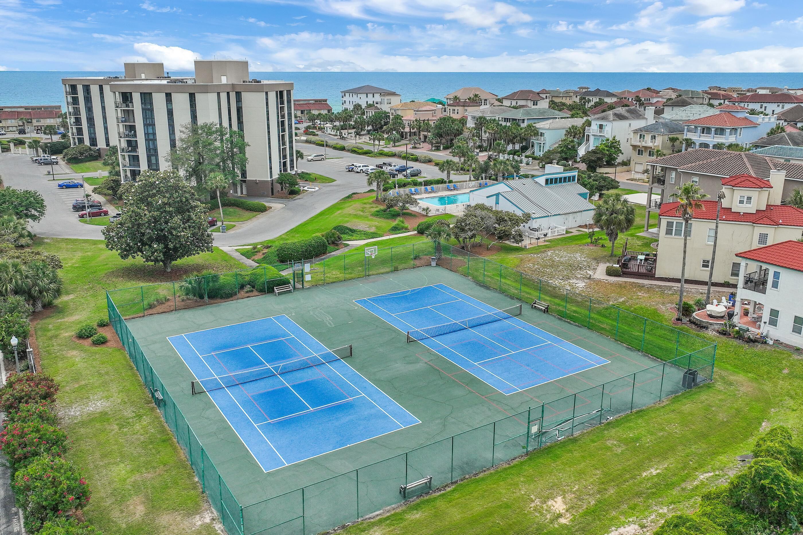 Una pista de tennis envoltada d'edificis residencials, una zona d'aparcament i vista a l'oceà.