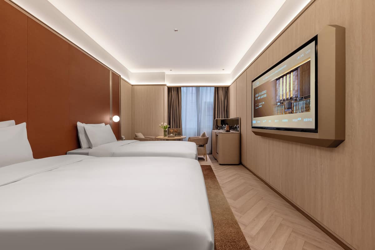 A hotel room with two beds, a TV mounted on the wall, a small desk, and a window with curtains.