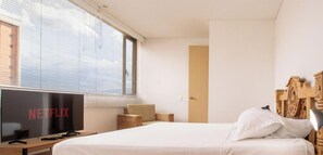A hotel room with a bed, a TV, a wooden headboard, a window with blinds, and a door. - Luxury 3 Bedroom Apartment in Provence (Medellín)