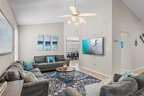 A living room with a ceiling fan, grey sofas, a coffee table, and a TV mounted on the wall. - Bamba's Beach House (Destin)
