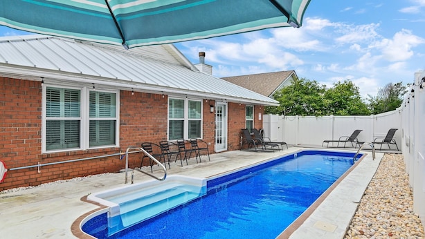 A rectangular swimming pool with a white deck, surrounded by a brick house with a white roof, and a covered patio area with chairs. - Bamba's Beach House (Destin)