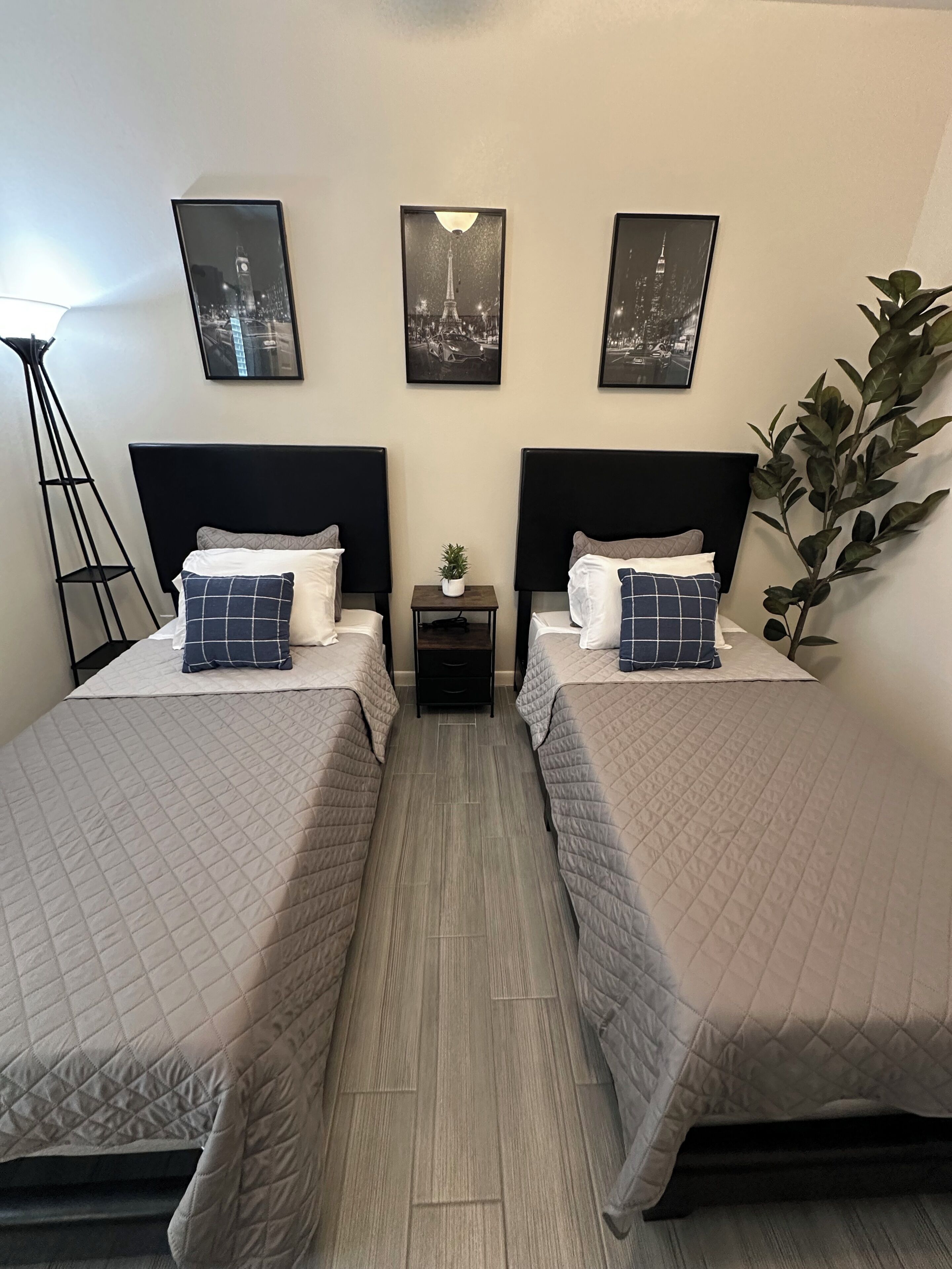 Een hotelkamer met twee bedden, een kleine tafel, een vloerlamp en drie ingelijste foto’s aan de muur.