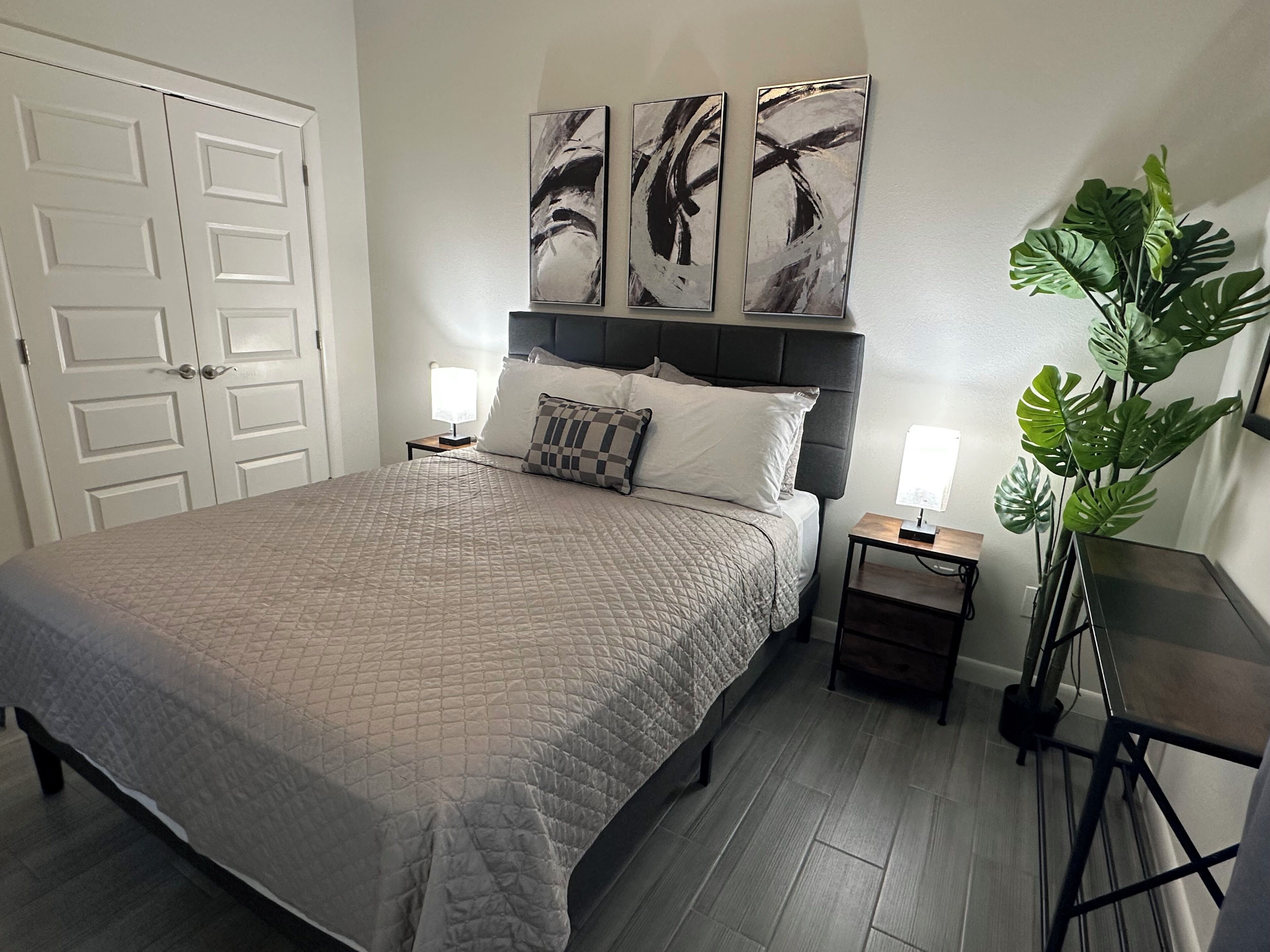 A bedroom with a bed, two bedside tables with lamps, a large potted plant, and abstract art on the wall.