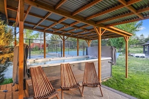 A covered outdoor hot tub area with wooden chairs and a pool in the background.