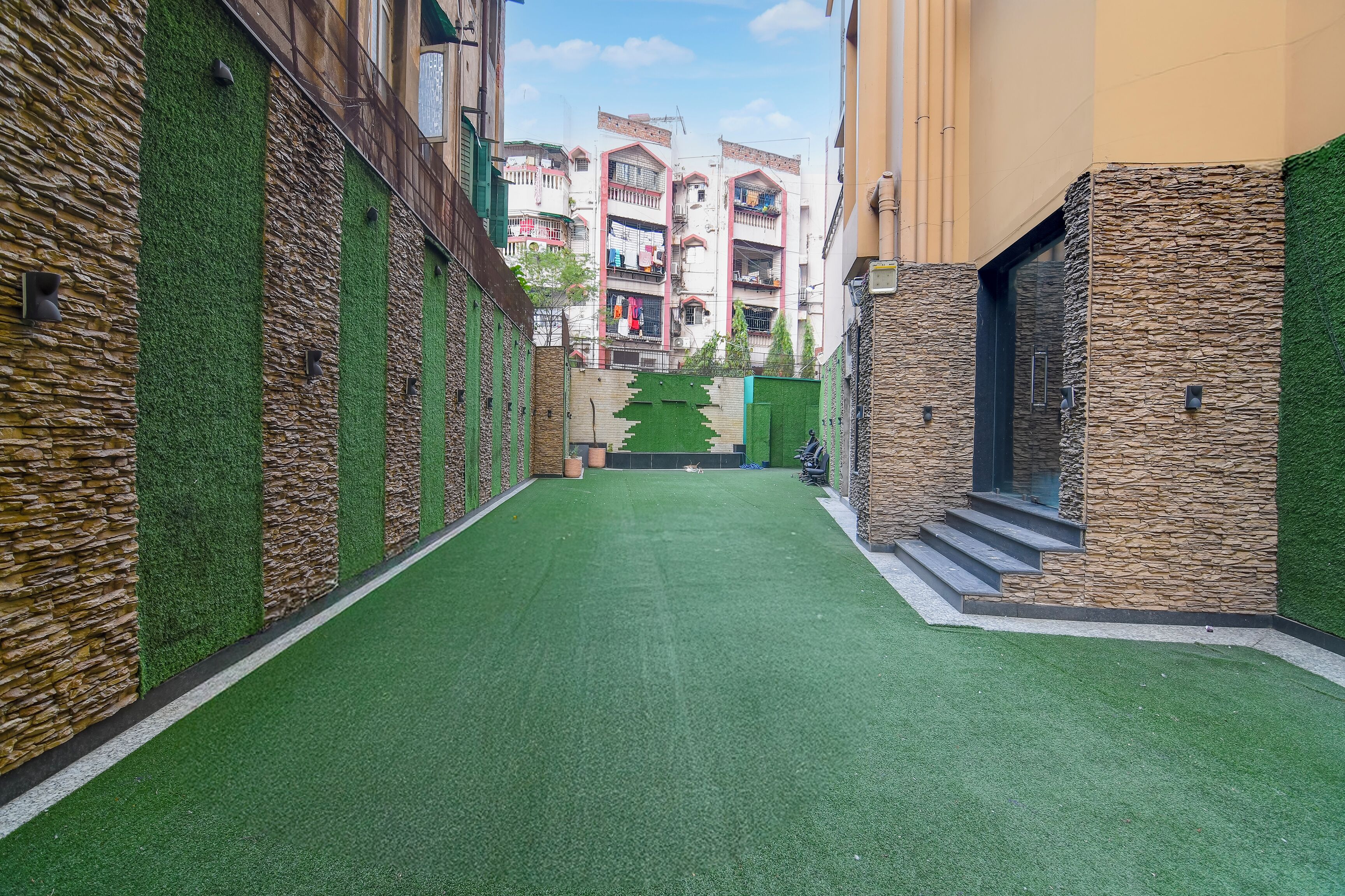 A narrow alleyway with artificial green turf, stone walls, and a view of multi-story buildings in the background.