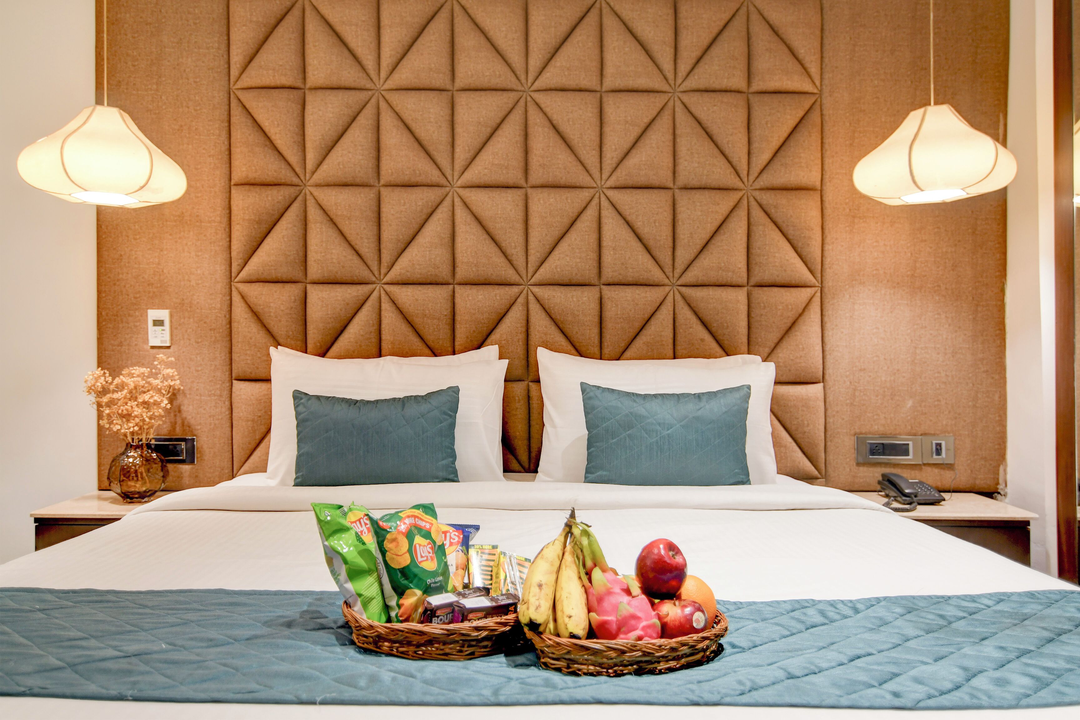 A hotel room with a large bed, two pillows, a nightstand with a phone, and two baskets of fruit and snacks on the bed.