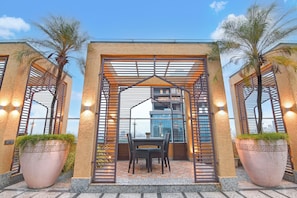 A covered outdoor seating area with a table and chairs, surrounded by large planters and a modern building in the background.